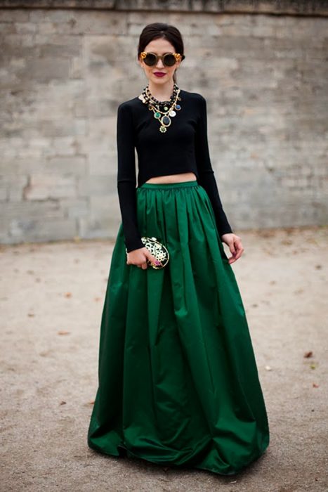 Menina com cabelo para cima usando óculos redondos, colar grande, blusa preta de mangas compridas e uma saia longa de cetim verde esmeralda