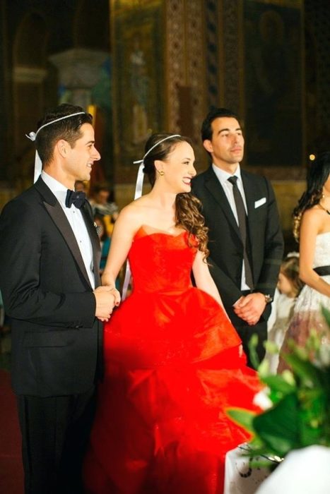 Um casal de noivos e seu padrinho em frente ao altar, ela usa um vestido vermelho sem alças e um laço branco na cabeça