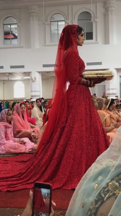 Uma noiva hindu em um vestido vermelho caminha pelo corredor de um templo cheio de pessoas sentadas no chão