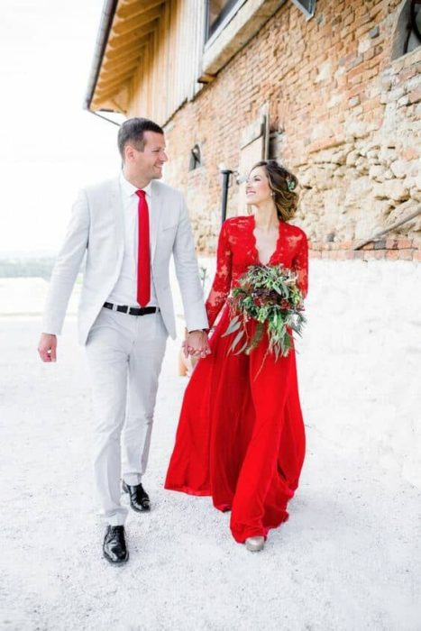 Um casal caminha de mãos dadas para a lateral de um prédio de pedra, ele em um terno cinza e gravata vermelha e ela em um vestido vermelho e com seu buquê