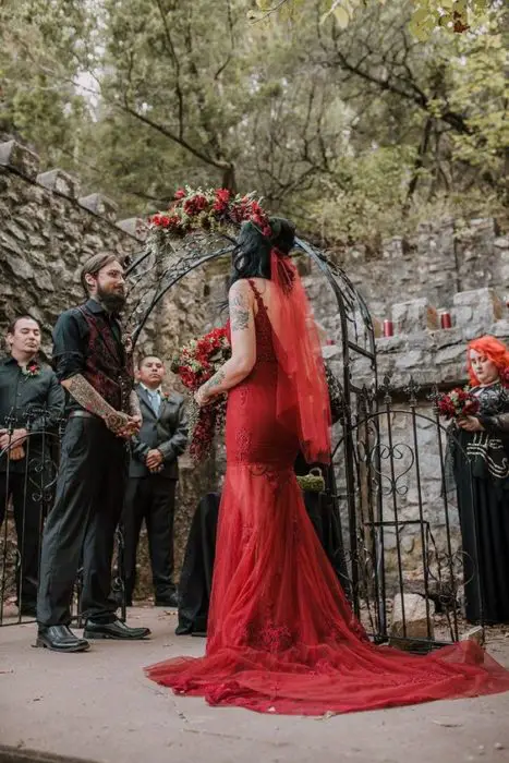um casal de noivos vai se casar em um jardim, ela em um vestido vermelho justo e um véu vermelho