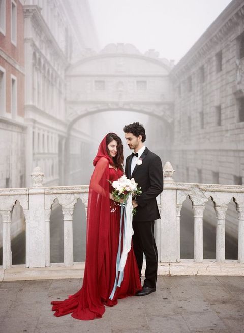 um casal de noivos posa em uma ponte do canal, ela em um vestido vermelho e um capuz da mesma cor