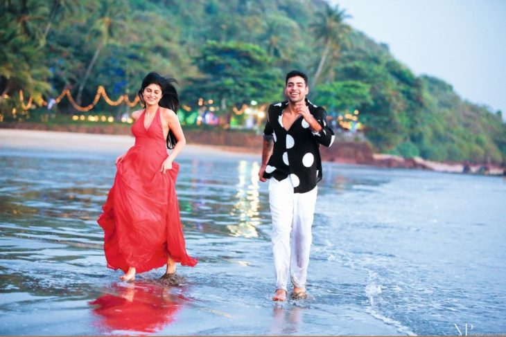 um casal está correndo pela praia, ele de camisa preta com calça branca e branca e ela de vestido vermelho