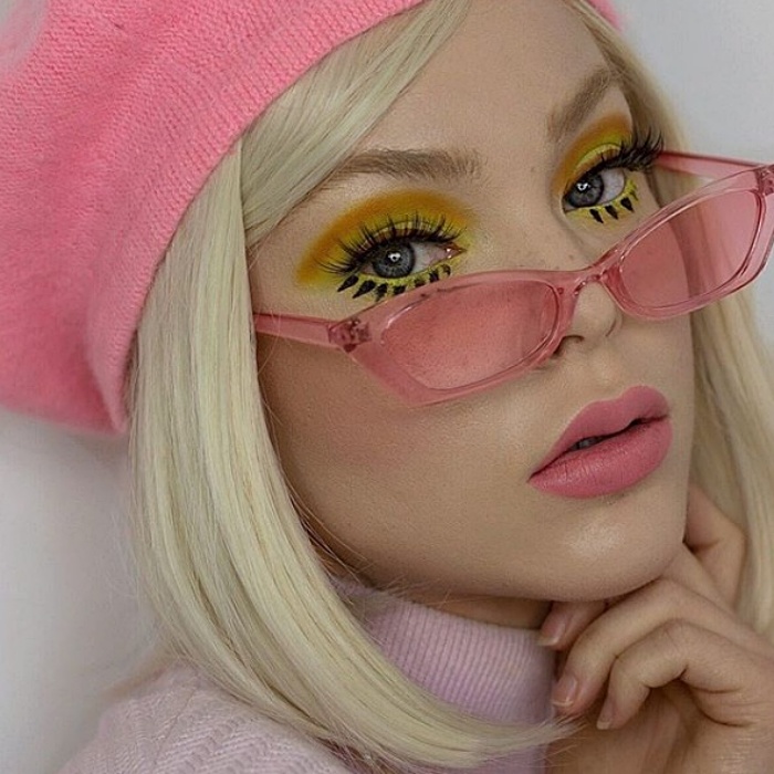 loira usando uma boina rosa, óculos de sol rosa, amarelo com maquiagem verde, delineador preto e batom rosa