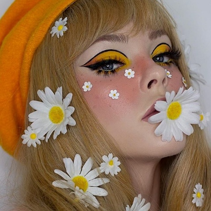 Menina loira de cabelos compridos usando margaridas florais no cabelo, com maquiagem de sombra amarela, forro preto, blush rosa e boina laranja