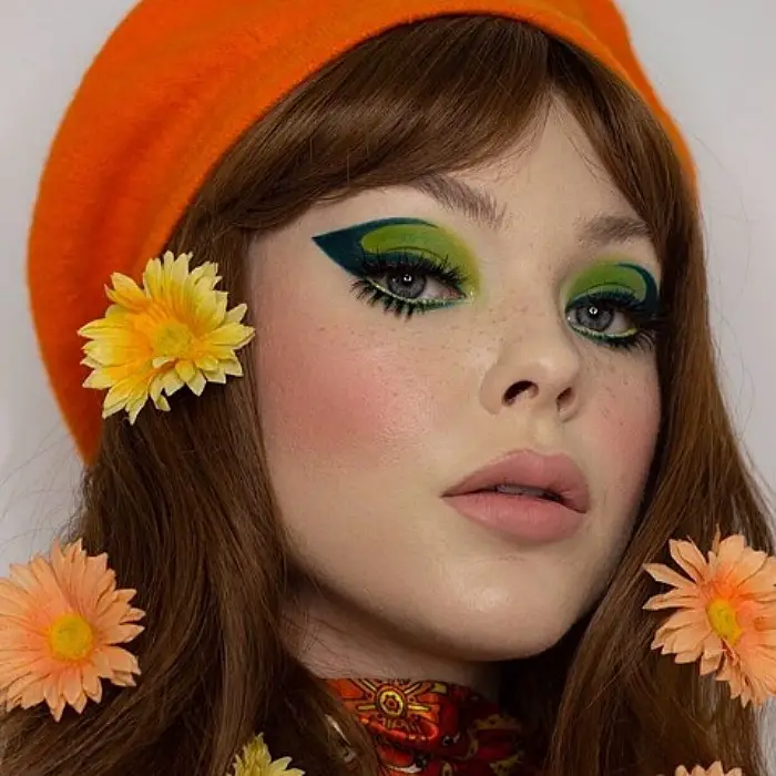 menina com cabelos castanhos compridos usando maquiagem com sombra verde escuro com contornos de flores laranja no cabelo